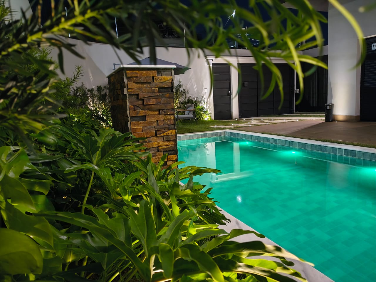 Piscina iluminada em turquesa vista por entre a vegetação à noite